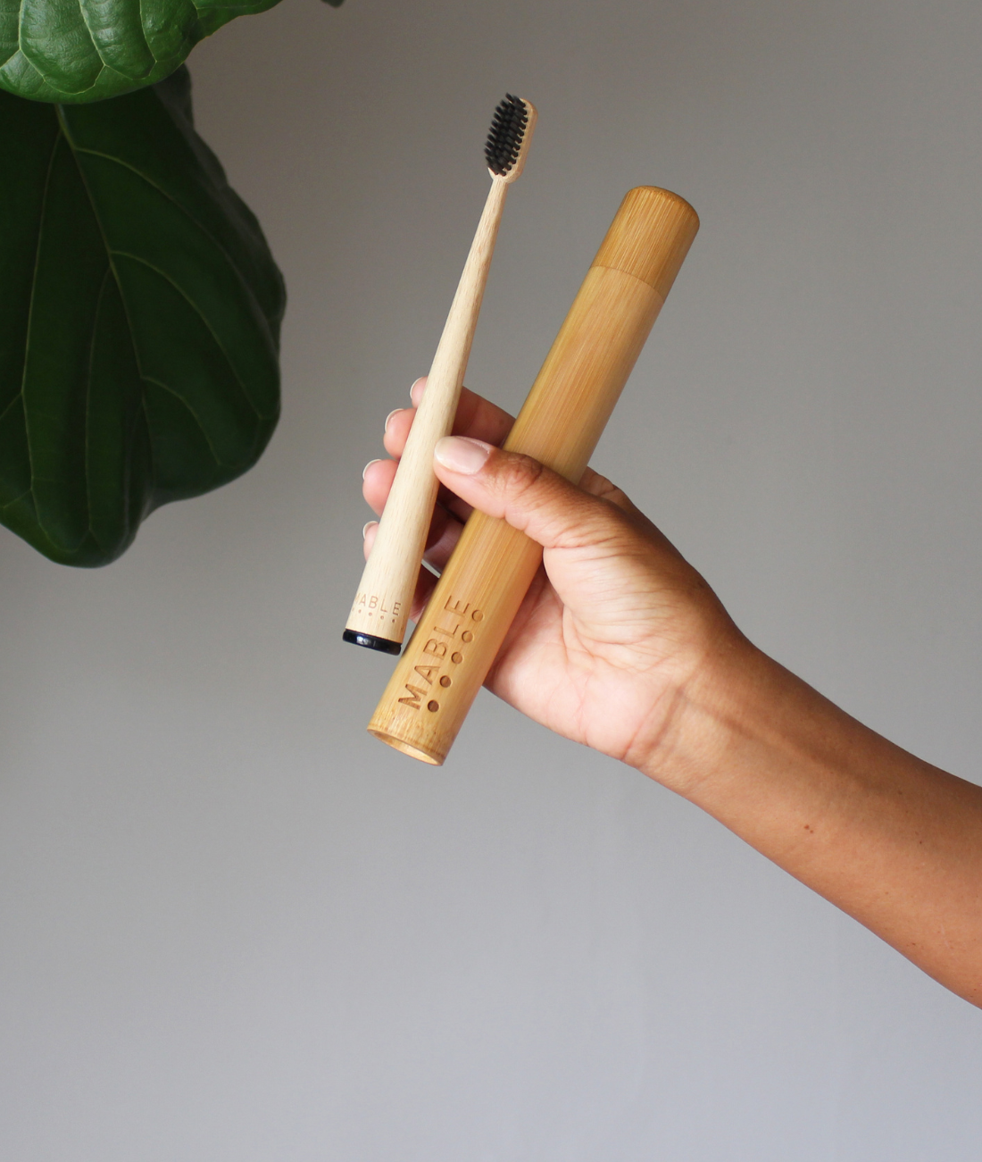 MABLE sustainable toothbrush holder with bamboo brush being held by a hand a neutral background with a bit of plant greenery showing