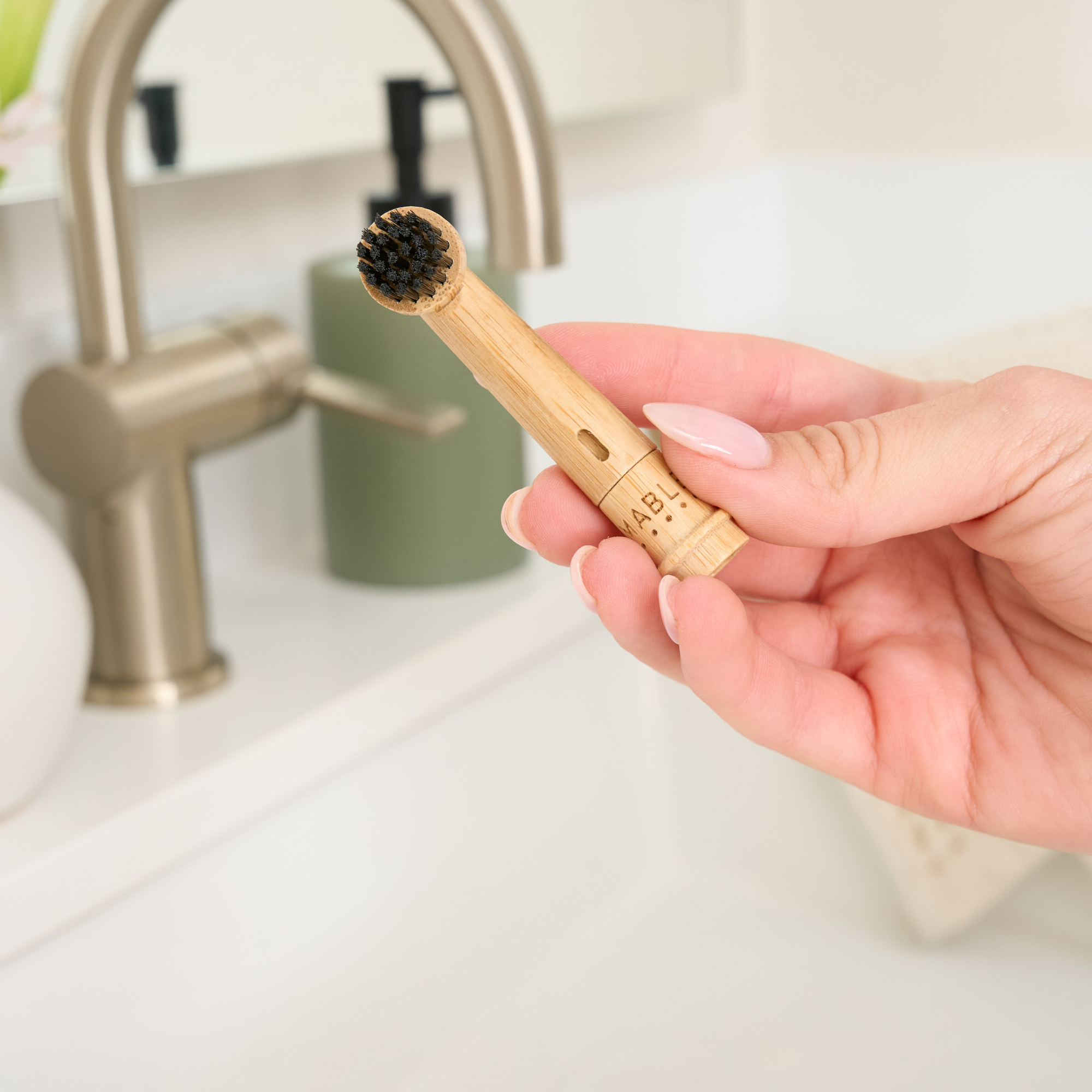 Hand holding a bamboo toothbrush head by MABLE for an Oral-B electric head with black bristles in a bathroom setting.