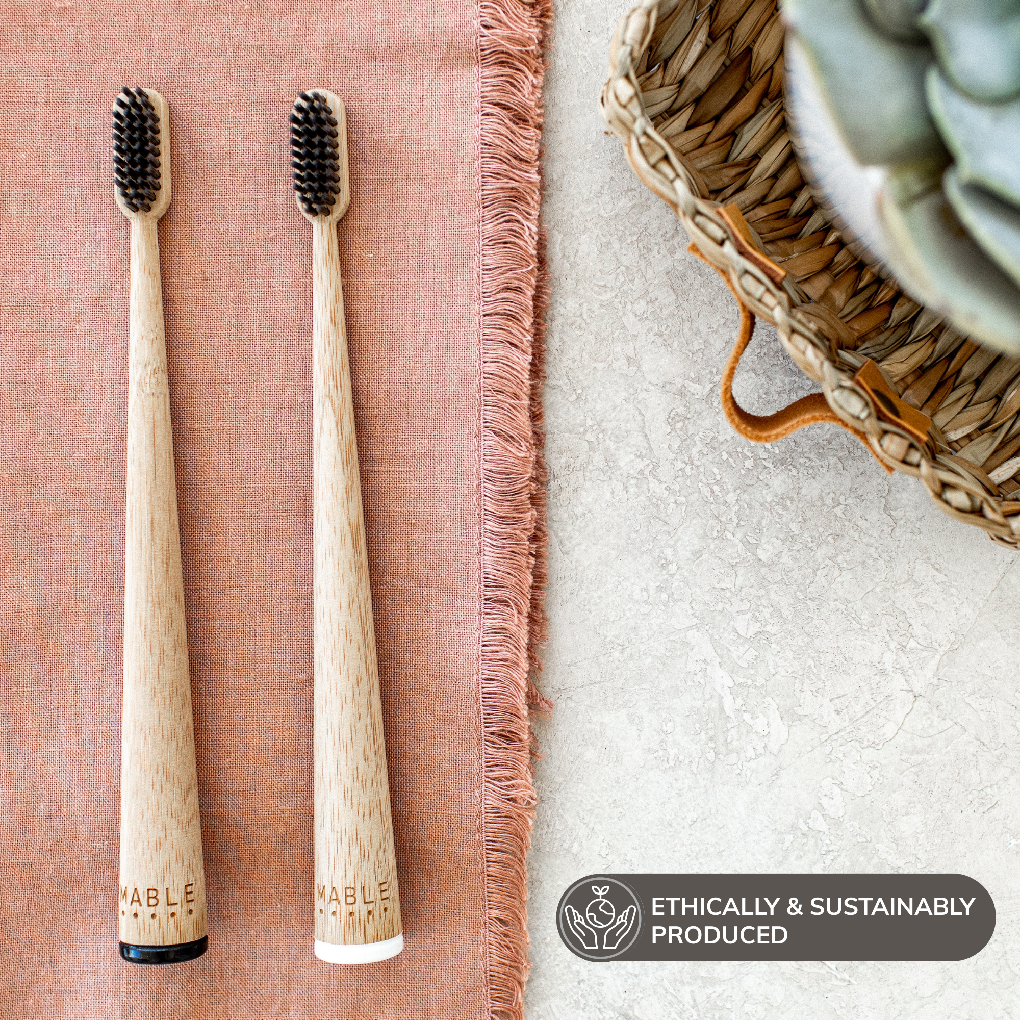 Two MABLE wooden toothbrushes with soft black bristles on a textured surface, accompanied by a basket and plant, badge saying ethically and sustainably produced