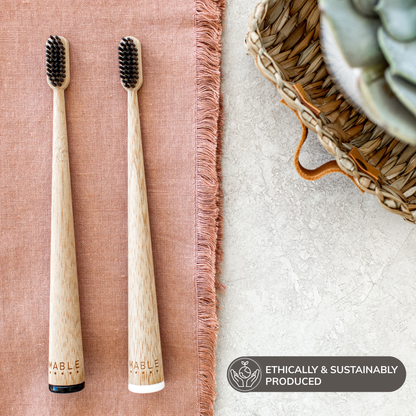Two MABLE wooden toothbrushes with soft black bristles on a textured surface, accompanied by a basket and plant, badge saying ethically and sustainably produced