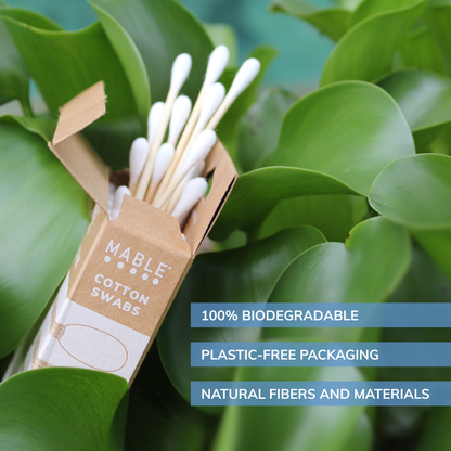 Mable cotton swabs in a cardboard box with green leaves in the background words reading 100% biodegradable, plastic-free packaging and natural fibers and materials