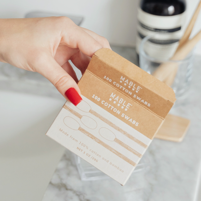 Hand holding a box of Mable Cotton Swabs on a marble surface.