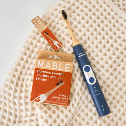 A Mable bamboo electric toothbrush head  on a dark blue Sonicare handle, shown next to its orange and kraft paper packaging box and a single replacement head, all arranged on a cream-colored waffle-weave towel.