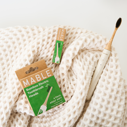 A Mable bamboo electric toothbrush head on a Philips One model with a cream handle and black plant-based bristles, resting on a textured cream waffle-knit blanket next to its green and kraft paper plastic-free packaging.