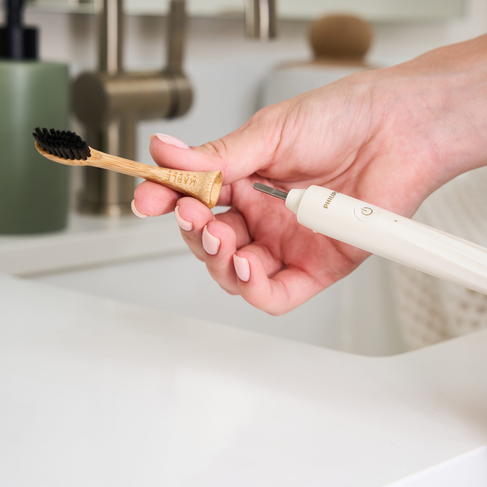 A hand holds a MABLE bamboo toothbrush head with black castor-oil  bristles next to a white Philips One electric toothbrush with a metal attachment, set in a bright bathroom sink area.