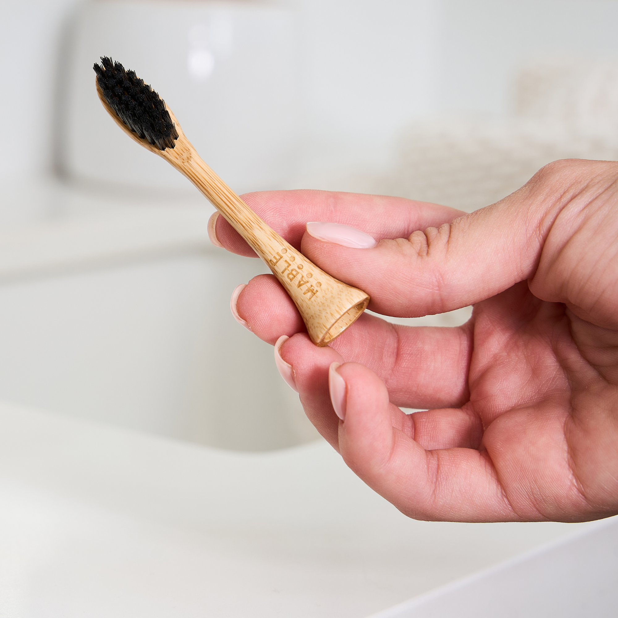 A hand holding a MABLE bamboo toothbrush head compatible with a Philips One electric toothbrush with black bristles, with a blurred bathroom sink background.